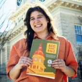 Artist Sam Watson smiles while holding a copy of the 2025 ACC from A to Z Guide while standing in front of City Hall