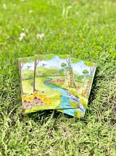 Picture of commemorative seed packets laying on grass. Seed packages has a watercolor style picture of a park with "Restore Our River, Dudley Park" written in the center in cursive