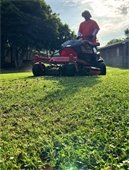 Crew member on an electric mower drives arcoss bright green grass with a building and a blue sky in the background